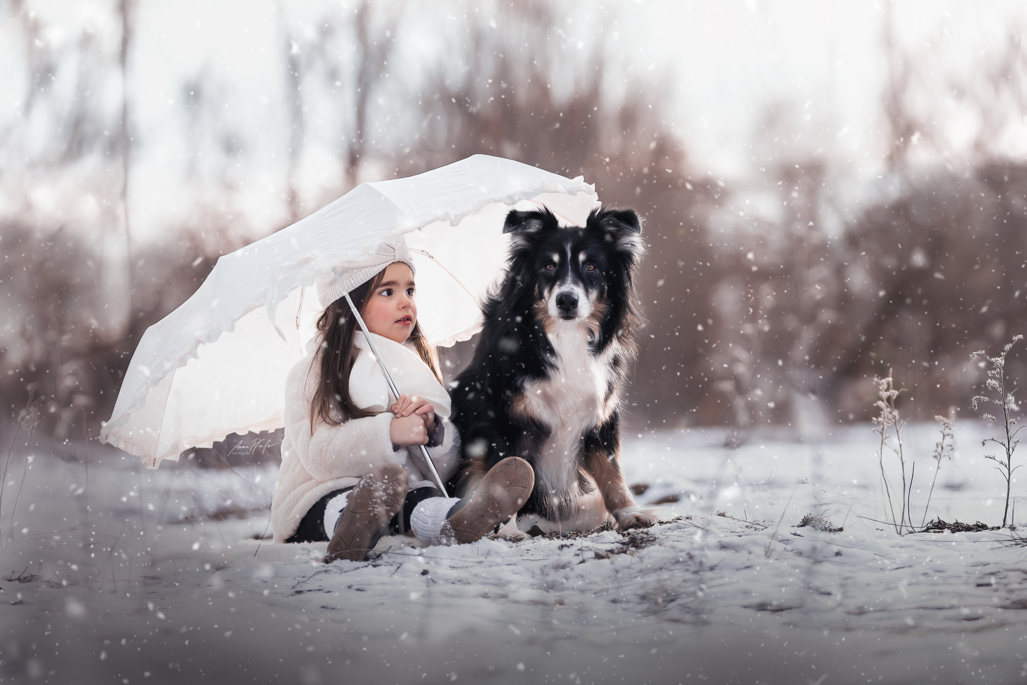 Kleines Mädchen und ihr Hund sitzen gemeinsam unter einem Schirm im Schnee